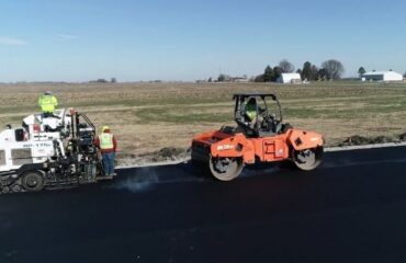 A paver and orange roller place a black asphalt mat beside fields at the BioCentury Research Farm.