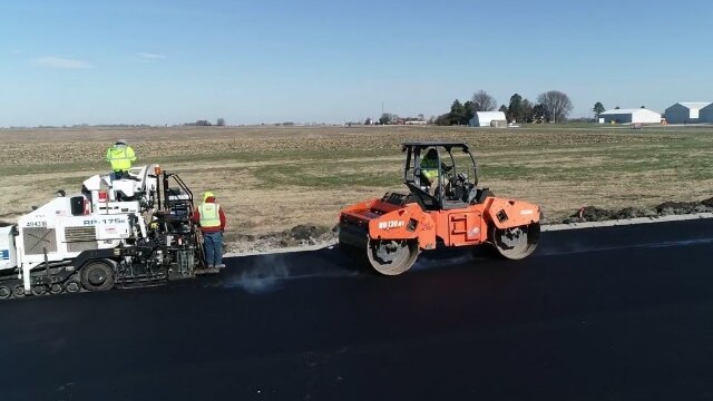 Boone, Iowa: 18% RAP and Polymer Performance with BioMAG™ A paver and orange roller place a black asphalt mat beside fields at the BioCentury Research Farm.