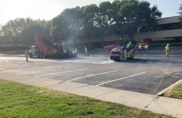 A paver, red haul truck, roller, and crew work in the Indiana Soybean Alliance parking lot.