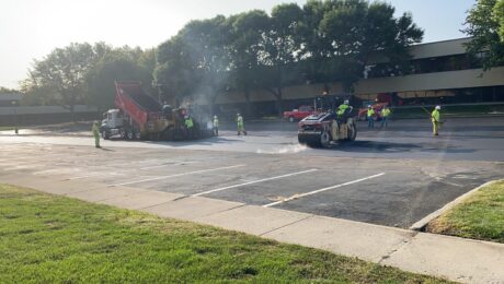 A paver, red haul truck, roller, and crew work in the Indiana Soybean Alliance parking lot.