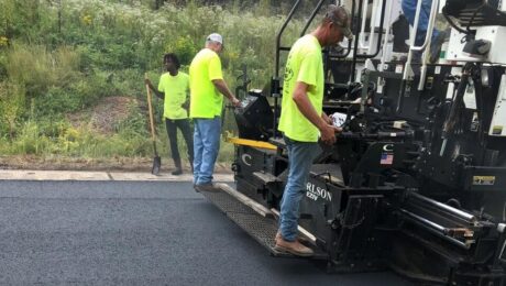 Three crew members stand on a paver placing a fresh asphalt mat at the NCAT Test Track.