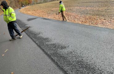Two crew members rake fresh asphalt on a rural road beside a harvested field.