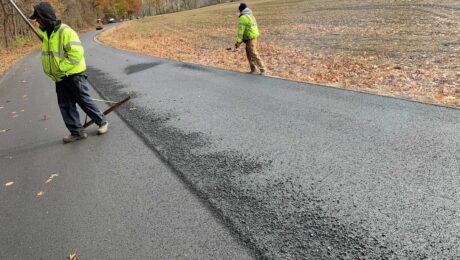 Two crew members rake fresh asphalt on a rural road beside a harvested field.