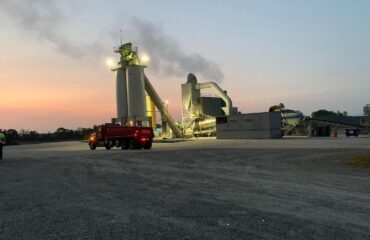 An asphalt plant operates at dusk with a red haul truck in front.