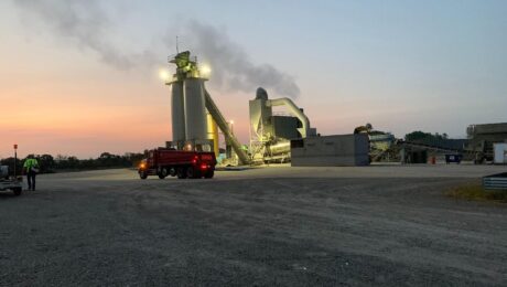 An asphalt plant operates at dusk with a red haul truck in front.