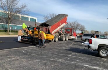 University Park, Illinois: 600 Tons of SIP-1111™ High-Recycled-Content Asphalt A paver, red haul truck, and crew place asphalt in a parking lot beside an office building.