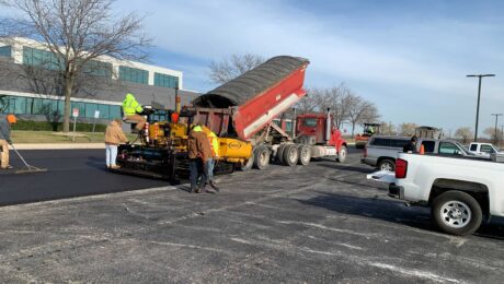 A paver, red haul truck, and crew place asphalt in a parking lot beside an office building.